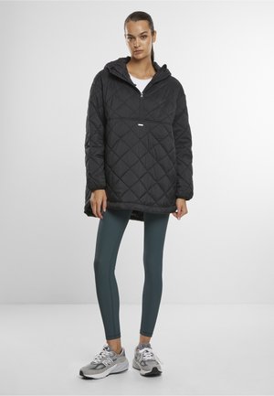 Vrouw die een zwarte doorgestikte pulloverjas met kap draagt, donkergroene leggings en grijze en witte sneakers, staand tegen een effen achtergrond.