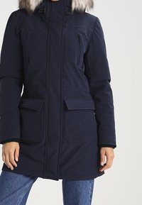 Marinblå vinterparka med huva dekorera med päls, två framfickor, dragkedjestängning och elastiska manschetter för värme och skydd mot kallt väder.