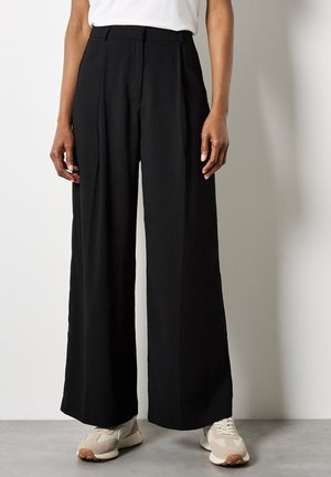 Schwarze, weite Hose mit vorderen Falten, hoher Taille und glattem Stoff, kombiniert mit hellen Sportschuhen auf einem grauen Boden.