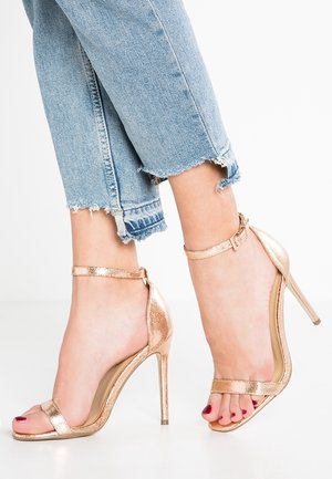 Des pieds chaussés de sandales à talons hauts métalliques rose gold avec des brides à la cheville, associées à un jean en denim bleu clair avec un ourlet effiloché.