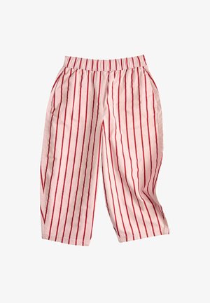 Lyshåret barn iført en rød knaplukket cardigan, hvide bukser med lodrette røde striber og beige sko, stående med hænderne i lommerne.