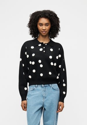 Woman with curly hair wearing a black long-sleeve polka dot sweater and light blue high-waisted jeans, standing against a plain background.