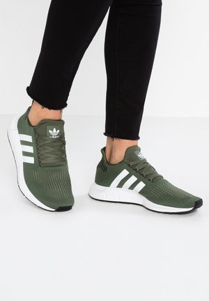 Personne portant des baskets Adidas vert olive avec des bandes blanches et un pantalon noir longueur cheville sur un sol blanc.