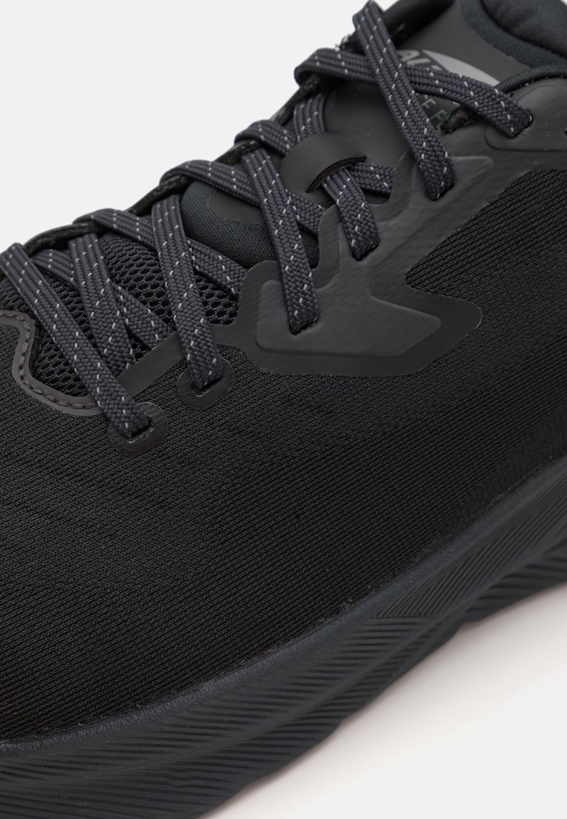 Zapatilla deportiva negra con un upper de tela texturizada, secciones de malla y acentos sintéticos. Los cordones y la suela de goma también son de color negro.