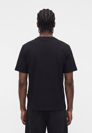 Osoba s pletenými vlasmi viditeľná zozadu, oblečená v jednoduchom čiernom tričku s krátkym rukávom a čiernych nohaviciach na bielom pozadí.