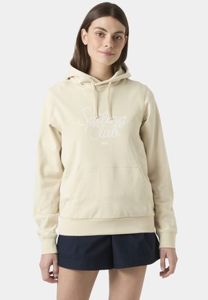 Jonge vrouw die een beige hoodie draagt met de tekst "Sailing Club" en donkerblauwe korte broek, staand tegen een effen lichte achtergrond.