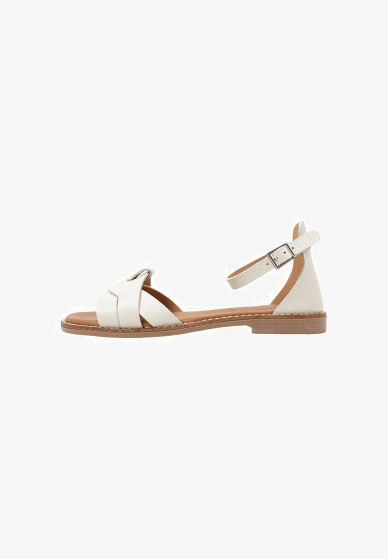 Sandale en cuir blanc avec un design en croix, orteils découverts, sangle de cheville réglable et semelle plate marron. Le matériau texturé ajoute des détails.