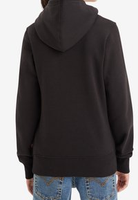 Černá mikina s kapucí a jemnou texturou, s klokankovou kapsou a žebrovanými manžetami. Nosič má na sobě modré džínové kalhoty, zobrazený pohled zezadu.