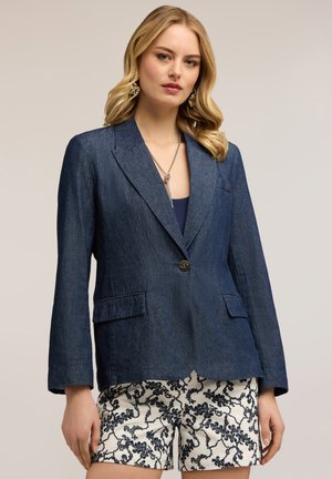 Blondynka w ciemnoniebieskiej marynarce na jeden guzik, granatowym topie i białych szortach z czarnym wzorem kwiatowym, pozująca na jednolitym tle.