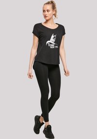 Svart T-skjorte med korte rullede ermer som har et hvitt grafisk bilde av en karakter og tekst. Kombinert med svarte leggings og sneakers.