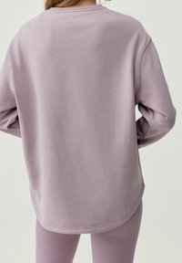 Vaaleanlaventelin värinen neule, jossa on pyöreä pääntie ja pitkät hihat. Sujuva pinta, rento istuvuus ja hieman kaareva helma takana.