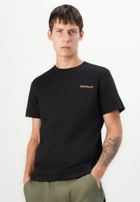 Zwarte katoenen t-shirt met korte mouwen, ronde hals en oranje Timberland-logo op de borst. Model draagt het met olijfgroene broeken.