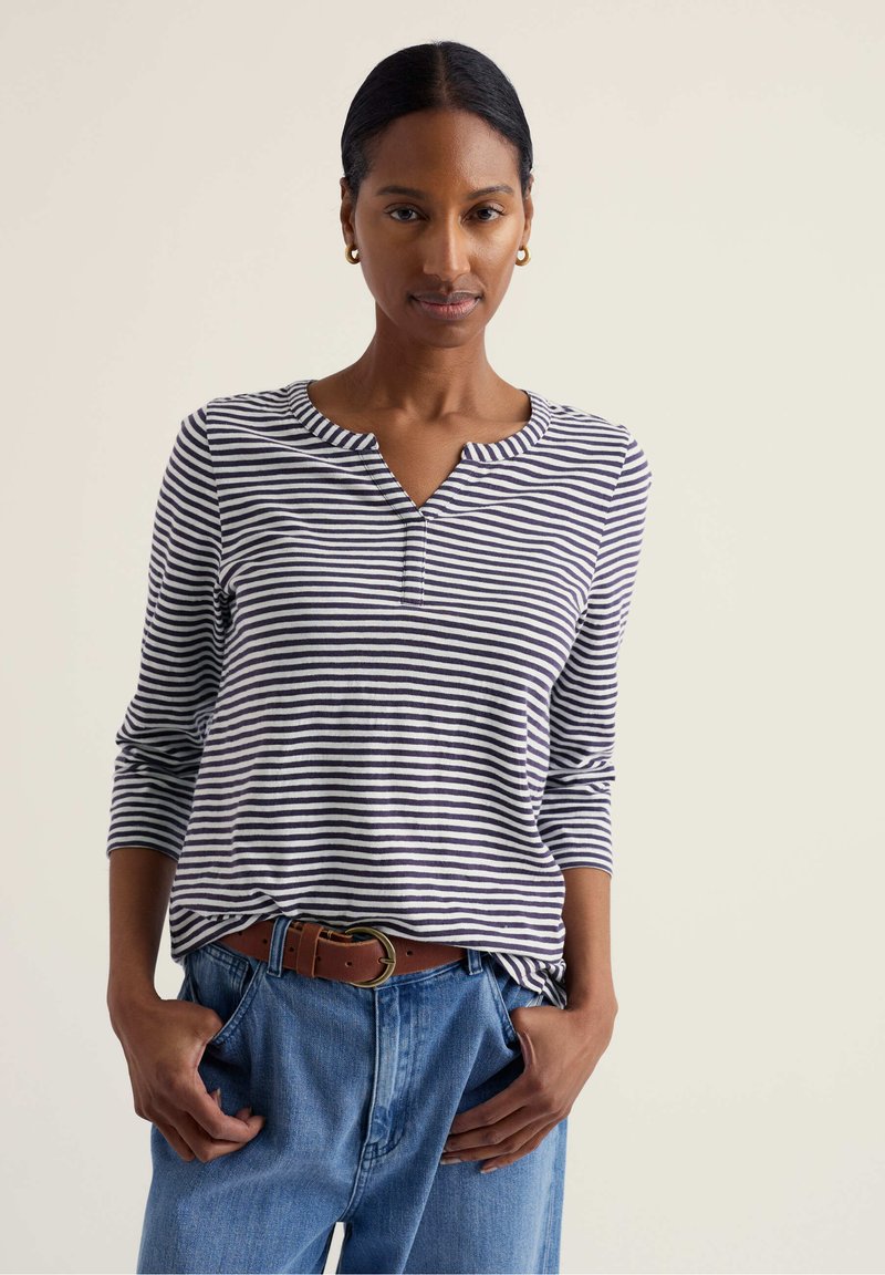 Vrouw met gouden ronde oorbellen, een marineblauw-wit gestreept shirt met lange mouwen, een bruine riem en een blauwe spijkerbroek met handen in de zakken.