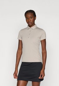 Beige polo shirt med krave, korte ærmer og knaplukning. Har et subtilt logo på kraven og tekst på ærmet. Sammen med et sort skørt.