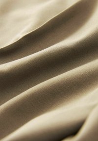 Tessuto beige con una texture liscia, caratterizzato da morbide pieghe fluenti che creano onde delicate e una leggera lucentezza in diverse condizioni di luce.