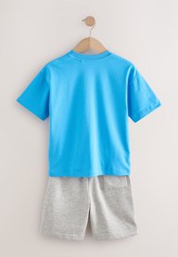 Tricou albastru cu mâneci scurte și guler rotund, asortat cu pantaloni scurți gri care au două buzunare în spate, agățate pe un umeraș din lemn pe un fundal deschis.