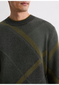Pull en vert foncé avec un tricot texturé et un motif en losanges. Il présente un col à côtes et des rayures d'accent jaune.