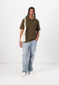 Polo shirt texturé vert olive, jean décontracté bleu clair, et baskets blanches. Modèle portant un sac fourre-tout beige.