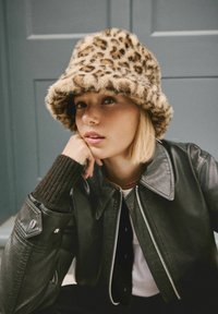 Leopard print faux fur bucket hat with a soft texture; worn with a black leather jacket featuring a silver zipper and ribbed sweater cuffs.