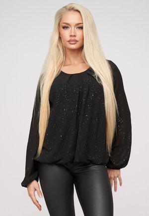 Femme blonde portant un haut noir à manches longues avec de petits détails scintillants et un pantalon en cuir noir, debout devant un fond uni.