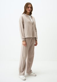Beige zip-up hoodie met een zachte textuur, gecombineerd met bijpassende wijde broek, beide met een ontspannen pasvorm en ribgebreide manchetten. Witte sneakers.