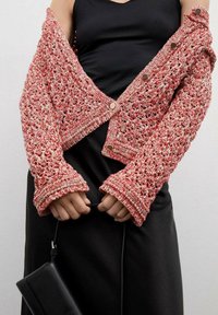 Pletený kardigán s otevřenou vazbou v růžové a bílé barvě, s cropped designem, zapínáním na knoflíky a širokými manžetami, v kombinaci s černými šaty a kabelkou.