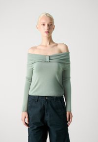 Off-shoulder ljusgrön topp med samlad tyg vid halsringningen, i kombination med mörka, löst sittande cargobyxor. Slät textur.