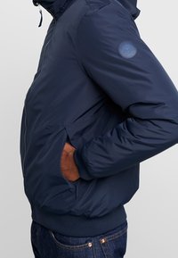 Navy blue bomber jacket, smooth texture, zippered front, side pockets, ribbed cuffs, logo patch on the upper sleeve, slightly padded design.