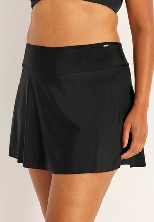 Kobieta nosząca czarną spódnicę kąpielową z wysokim stanem, z małą metalową etykietą na pasku, stoi z prawą ręką w spoczynku.