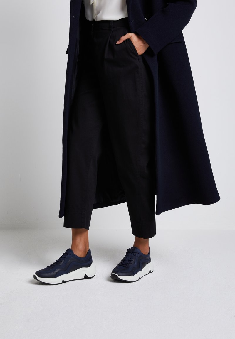 Personne portant un pantalon noir court, un long manteau bleu marine et des baskets bleu marine à semelles blanches, debout avec une main dans la poche sur un fond clair.