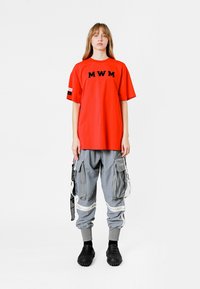 Camiseta roja con bordado "MWM", combinada con pantalones cargo grises que tienen múltiples bolsillos y acentos blancos. Unos zapatos negros texturizados completan el conjunto.