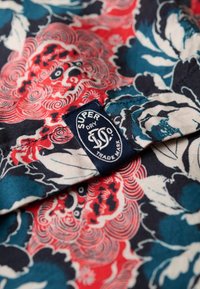 Kleurrijke katoenen stof met bloemenpatronen in rood, blauw en wit, met een blauw label waarop "SUPERDRY TRADE MARK" is gestikt.