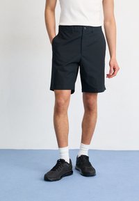 Man som står iklädd svarta shorts, vita strumpor, svarta sneakers och en vit ärmlös tröja mot en ljus bakgrund och blå golv.