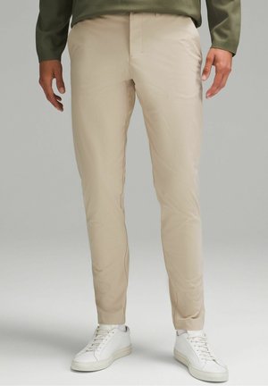 Mężczyzna ubrany w beżowe, dopasowane spodnie i białe trampki stoi na jasnoszarym tle.