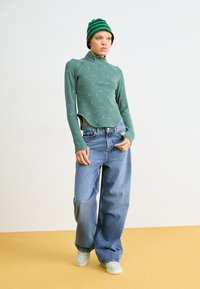 Grønn glidelås-langermet topp med teksturert mønster, kombinert med løse blå jeans og lysegrønne sko. Bakgrunnen er lys grå.