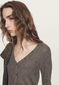 Šedý kardigan s výstřihem do V a knoflíky, vyrobený z měkké látky. Má mírně volný střih a texturovaný vzor podél límce a rukávů.