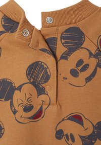 Brauner Stoff mit einem sich wiederholenden Mickey-Mouse-Muster, ausgestattet mit einem Rundhalsausschnitt und Druckknöpfen sowie strukturierten Akzenten am Kragen.
