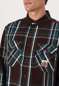 Brown, green, and white plaid flannel shirt with button-up front and two chest pockets. Features a tag with the "Element" logo.