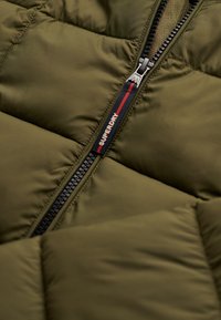 Section de doudoune vert olive avec texture matelassée, dotée d'une fermeture éclair noire avec des accents rouges et blancs ainsi que d'un logo "SUPERDRY".