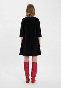 Robe noire en ligne A avec des manches trois-quarts, fabriquée en tissu doux. Associée à des bottes en cuir rouge montantes pour le contraste.