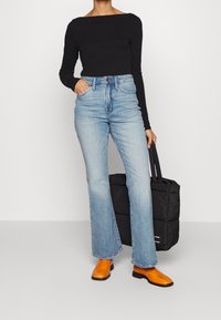 Top negro de manga larga; jeans acampanados azul claro; botas de cuero naranjas hasta el tobillo; sosteniendo una bolsa tote negra acolchada. Diseño simple y minimalista.