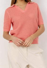 Nettimäinen lyhyt hiha pullover korallinpunaisena, jossa on V-pääntie ja kaulus, hienostuneella tekstuurikuviolla ja ribbaahelma. Yhdistetty kermaisiin housuihin.