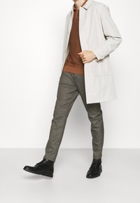 Cappotto leggero beige sopra una polo marrone, abbinato a pantaloni grigio a scacchi e stivali neri con lacci. Design semplice e su misura.