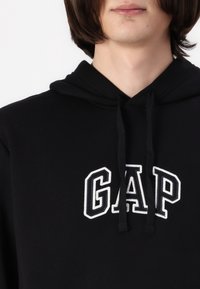 Haut à capuche noir avec une poche kangourou et un cordon de serrage. Présente un logo "GAP" brodé en blanc sur la poitrine. Tissu doux au toucher.