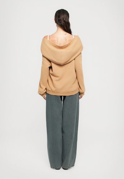 Femme avec de longs cheveux noirs, portant un hoodie beige à épaules dénudées, des bretelles de soutien-gorge blanches et un pantalon gris à jambes larges, standing de dos sur fond blanc.