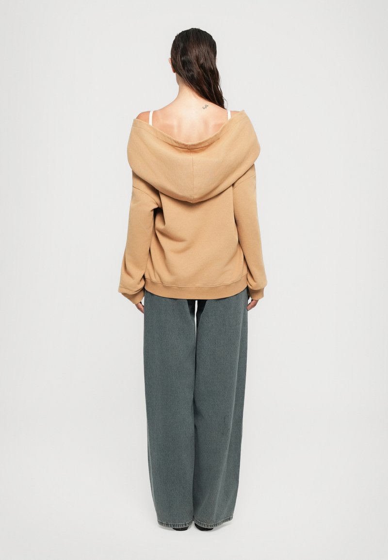 Kvinna med långt mörkt hår, klädd i en beige off-shoulder hoodie, vita bh-banor och vida grå byxor, stående vänd bort från kameran mot vit bakgrund.