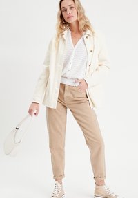 Light cream jacket, white eyelet blouse, beige corduroy pants, and tan high-top sneakers. Model holds a cream handbag.