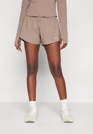 Osoba ubrana w beżowe sportowe spodenki Nike, pasujący beżowy longsleeve, białe skarpetki i jasne buty do biegania stojąca na białej podłodze.