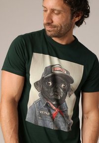 Groene katoenen t-shirt met een afbeelding van een hond in een spijkerjasje en pet, met rode accenten, belichamend een speelse, humoristische stijl.