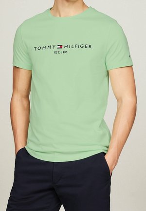 Homme portant un t-shirt Tommy Hilfiger vert clair avec logo et texte "Est. 1985", associé à un pantalon bleu marine foncé, les mains dans les poches.